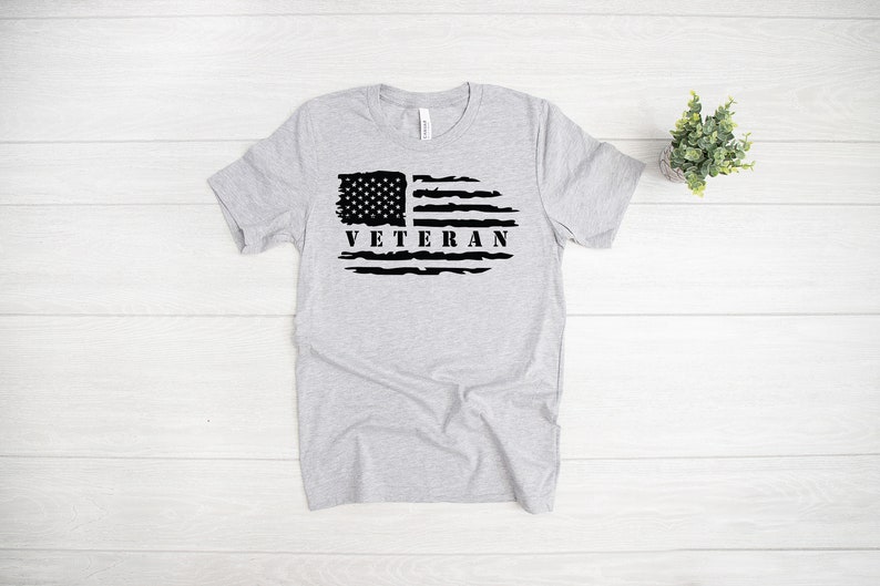 Może przedstawiać: Szary T-shirt z nadrukowaną czarną, przetartą flagą amerykańską i napisem "Veteran".