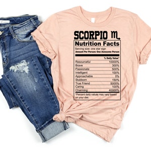 May include: A peach-colored t-shirt with the text "SCORPIO M. Nutrition Facts" and a table listing personality traits with percentages. The table includes traits like "Resourceful", "Brave", "Passionate", "Intelligent", "Approachable", "Stubborn", "True Friend", "Caring", and "Charming".