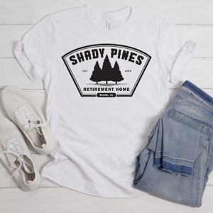 Może przedstawiać: Biała koszulka z czarnym nadrukiem graficznym, na kt&oacute;rym widnieje napis "Shady Pines Retirement Home" wraz z logo dw&oacute;ch sosen i tekstem "Est. 1985" oraz "Miami, FL".