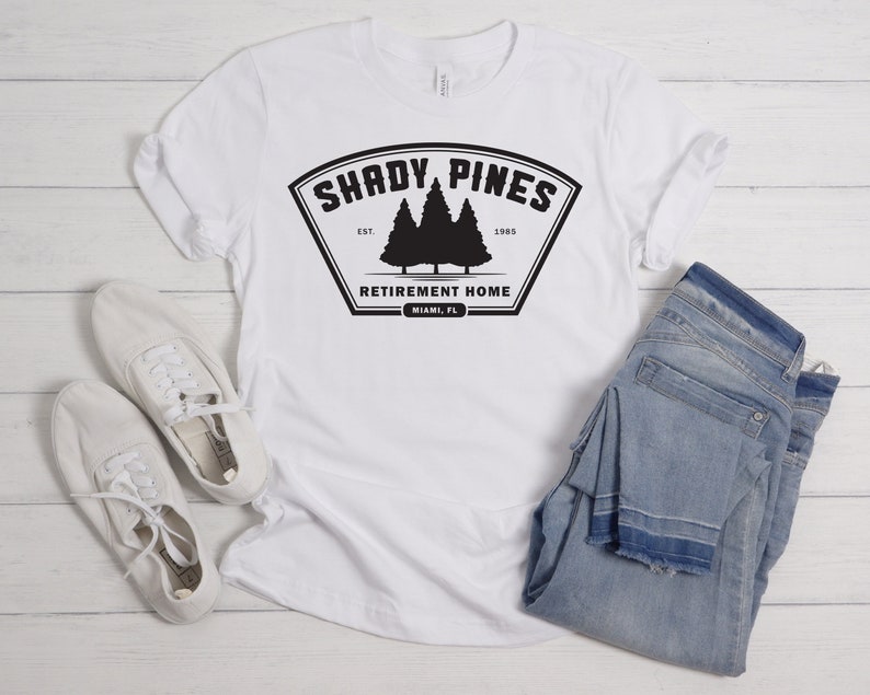 Może przedstawiać: Biała koszulka z czarnym nadrukiem graficznym, na kt&oacute;rym widnieje napis "Shady Pines Retirement Home" wraz z logo dw&oacute;ch sosen i tekstem "Est. 1985" oraz "Miami, FL".