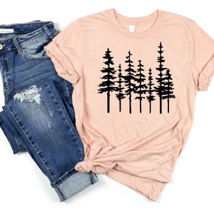 May include: A light pink t-shirt featuring a black graphic of several pine trees. The shirt is paired with blue denim jeans. The t-shirt has rolled-up sleeves and a knot at the bottom.