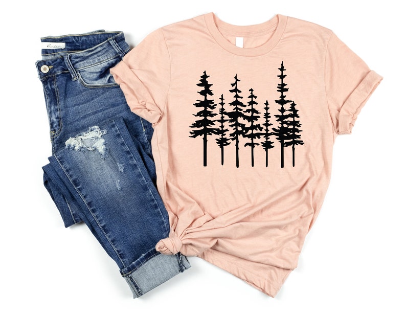 May include: A light pink t-shirt featuring a black graphic of several pine trees. The shirt is paired with blue denim jeans. The t-shirt has rolled-up sleeves and a knot at the bottom.