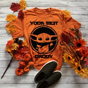 K&ouml;nnte beinhalten: Orangefarbenes T-Shirt mit einem Schwarz-Wei&szlig;-Bild eines Charakters aus dem Star Wars-Franchise, bekannt als Baby Yoda. Der Text "Yoda Best Daddy" ist auf dem Shirt aufgedruckt.