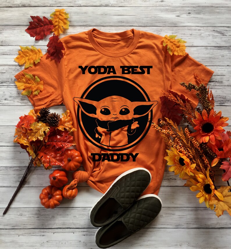 K&ouml;nnte beinhalten: Orangefarbenes T-Shirt mit einem Schwarz-Wei&szlig;-Bild eines Charakters aus dem Star Wars-Franchise, bekannt als Baby Yoda. Der Text "Yoda Best Daddy" ist auf dem Shirt aufgedruckt.