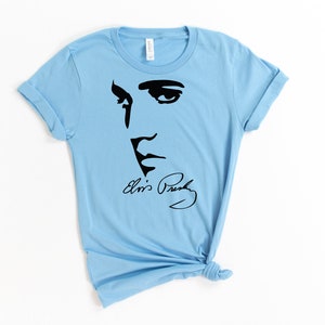 May include: Light blue t-shirt with a black silhouette of Elvis Presley's face and his signature "Elvis Presley".