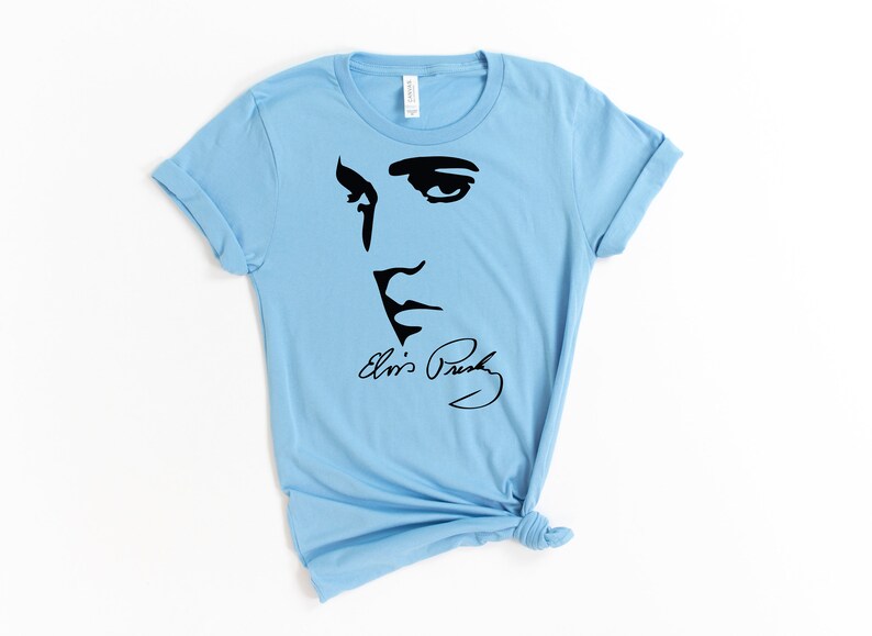 May include: Light blue t-shirt with a black silhouette of Elvis Presley's face and his signature "Elvis Presley".
