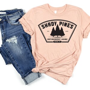 Może przedstawiać: R&oacute;żowy t-shirt z czarnym nadrukiem logo z napisem "Shady Pines Retirement Home" oraz tekstem "Est. 1985" i "Miami, FL".