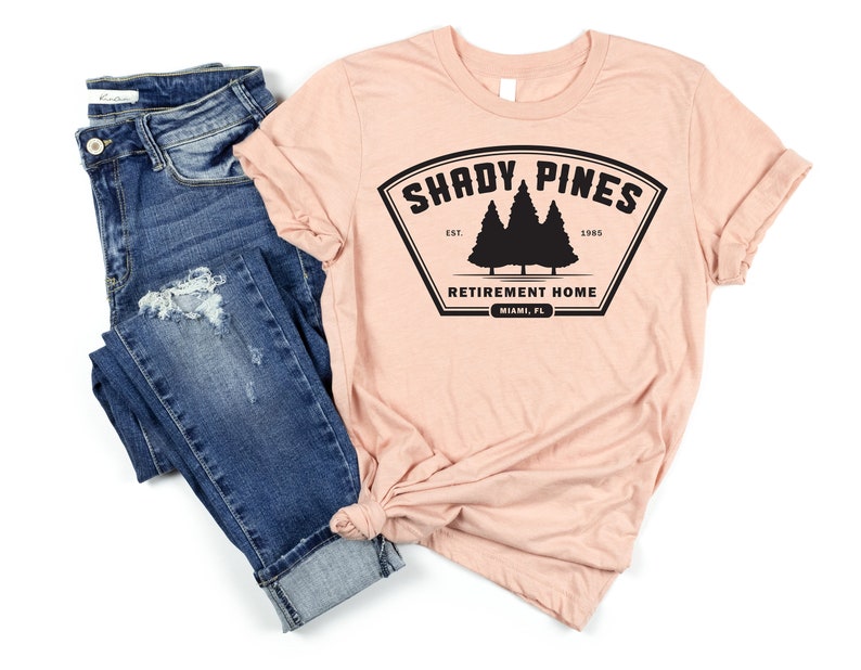 Może przedstawiać: R&oacute;żowy t-shirt z czarnym nadrukiem logo z napisem "Shady Pines Retirement Home" oraz tekstem "Est. 1985" i "Miami, FL".