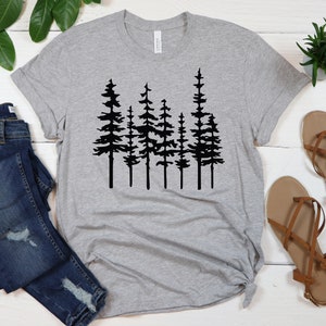 May include: A heather gray t-shirt featuring a black silhouette of a forest of pine trees. The shirt is paired with blue denim jeans and brown sandals. The shirt is made of a soft material.