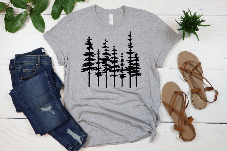 May include: A heather gray t-shirt featuring a black silhouette of a forest of pine trees. The shirt is paired with blue denim jeans and brown sandals. The shirt is made of a soft material.