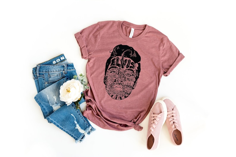 May include: A mauve t-shirt with a black graphic of Elvis Presley's face made up of words. The graphic includes the word "Elvis" across the forehead. The t-shirt is paired with blue jeans and pink sneakers.