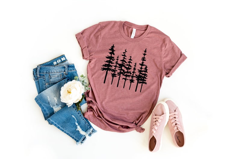 May include: Mauve-colored t-shirt with a black tree silhouette design. The image also includes blue denim jeans, a white flower, and pink sneakers. The t-shirt is knotted at the waist.