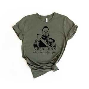 K&ouml;nnte beinhalten: Ein gr&uuml;nes T-Shirt mit einer schwarzen Grafik eines Mannes, der ein Messer h&auml;lt. Der Text "A real man will chase after you" ist unter der Grafik gedruckt.