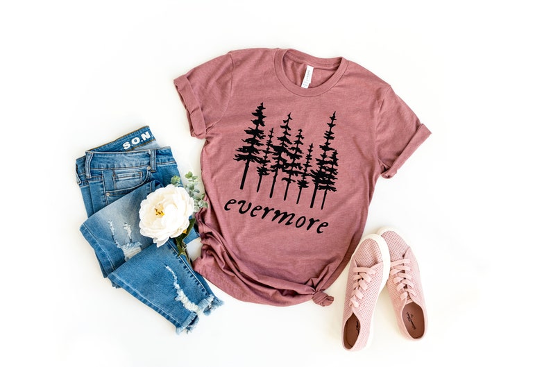 May include: A mauve-colored t-shirt with a black tree graphic and the word "evermore" in a script font. The shirt is paired with distressed blue jeans, pink sneakers, and a white flower on a white background.