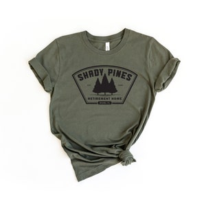 Może przedstawiać: Zielony t-shirt z czarnym nadrukiem, na kt&oacute;rym widnieje napis "Shady Pines Retirement Home" z logo trzech sosen.