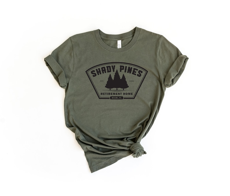 Może przedstawiać: Zielony t-shirt z czarnym nadrukiem, na kt&oacute;rym widnieje napis "Shady Pines Retirement Home" z logo trzech sosen.