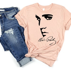 May include: A peach-colored t-shirt with a black silhouette of Elvis Presley's face and signature. The t-shirt is paired with blue jeans.