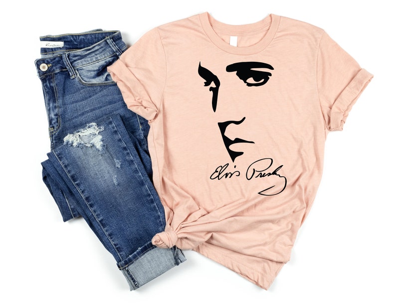 May include: A peach-colored t-shirt with a black silhouette of Elvis Presley's face and signature. The t-shirt is paired with blue jeans.