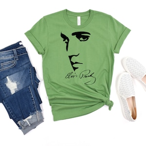 May include: A green t-shirt with a black silhouette of Elvis Presley's face and signature. The t-shirt is folded and displayed with a pair of blue jeans and white slip-on shoes.