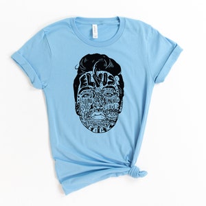 May include: Light blue t-shirt with a black and white graphic of Elvis Presley's face. The graphic is made up of words related to Elvis's music and career, including "Hound Dog", "Jailhouse Rock", and "Can't Help Falling in Love".