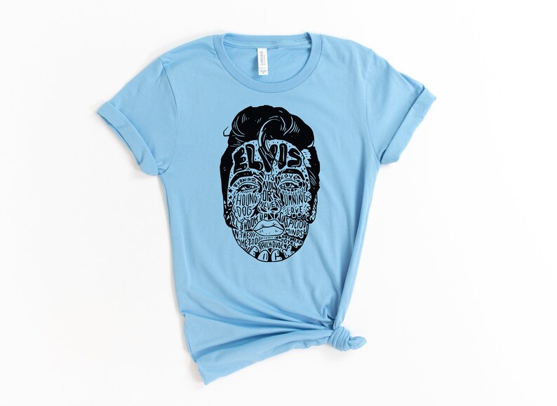 May include: Light blue t-shirt with a black and white graphic of Elvis Presley's face. The graphic is made up of words related to Elvis's music and career, including "Hound Dog", "Jailhouse Rock", and "Can't Help Falling in Love".
