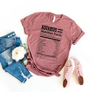 May include: A mauve t-shirt with a black graphic of a nutrition facts label for "Aquarius" with a wavy line symbol. The label lists traits like "Progressive", "Original", "Humanitarian", "Independent", "Analytical", "Rebellious", "Friendly", "Helping Hand", "Intelligent", "Stubborn", and "Creative".