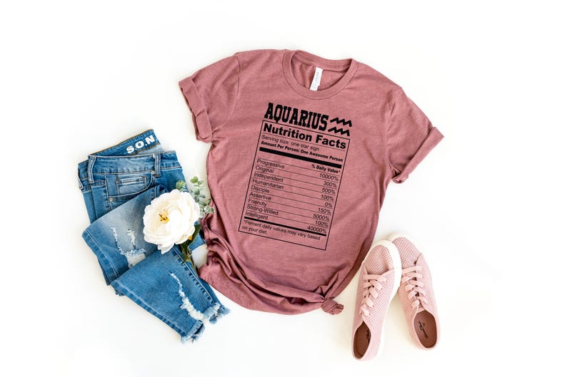 May include: A mauve t-shirt with a black graphic of a nutrition facts label for "Aquarius" with a wavy line symbol. The label lists traits like "Progressive", "Original", "Humanitarian", "Independent", "Analytical", "Rebellious", "Friendly", "Helping Hand", "Intelligent", "Stubborn", and "Creative".