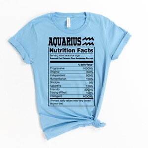 May include: A light blue t-shirt with a black graphic of a nutrition facts label for Aquarius. The label lists traits like "Progressive", "Original", "Independent", and "Intelligent" with percentages next to each trait.