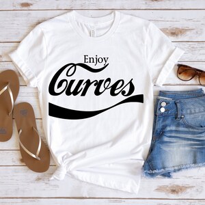 May include: White t-shirt with black text that says "Enjoy Curves" in a retro font style. The shirt is laid out on a wooden surface with a pair of brown flip flops, denim shorts, and sunglasses.