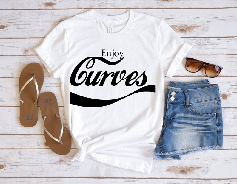 May include: White t-shirt with black text that says "Enjoy Curves" in a retro font style. The shirt is laid out on a wooden surface with a pair of brown flip flops, denim shorts, and sunglasses.