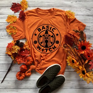 May include: Orange t-shirt featuring a black graphic of a witch wearing a hat and a coffee cup with the text "BASIC WITCH" around it. The shirt is laid out on a wooden surface with autumn leaves and pumpkins.