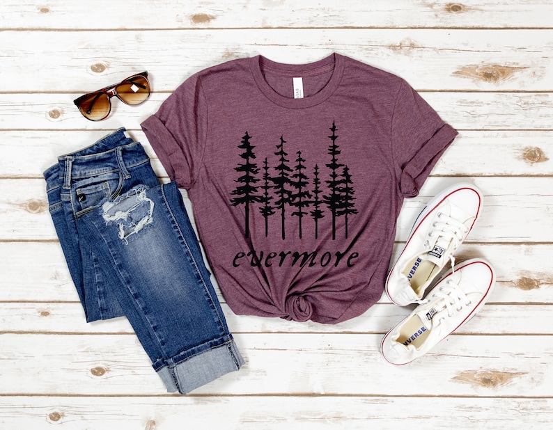 May include: A maroon t-shirt with a black tree graphic and the word "evermore" in a script font. The shirt is paired with blue jeans, white sneakers, and sunglasses. The items are arranged on a white wooden surface.