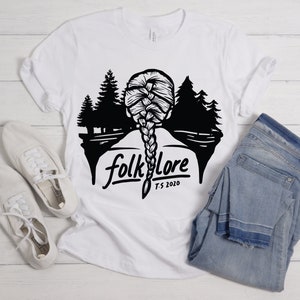 May include: White t-shirt with a black graphic featuring a braided hairstyle, trees, and the word "folklore" with "T.S. 2020" underneath. The shirt is paired with white sneakers and blue jeans.
