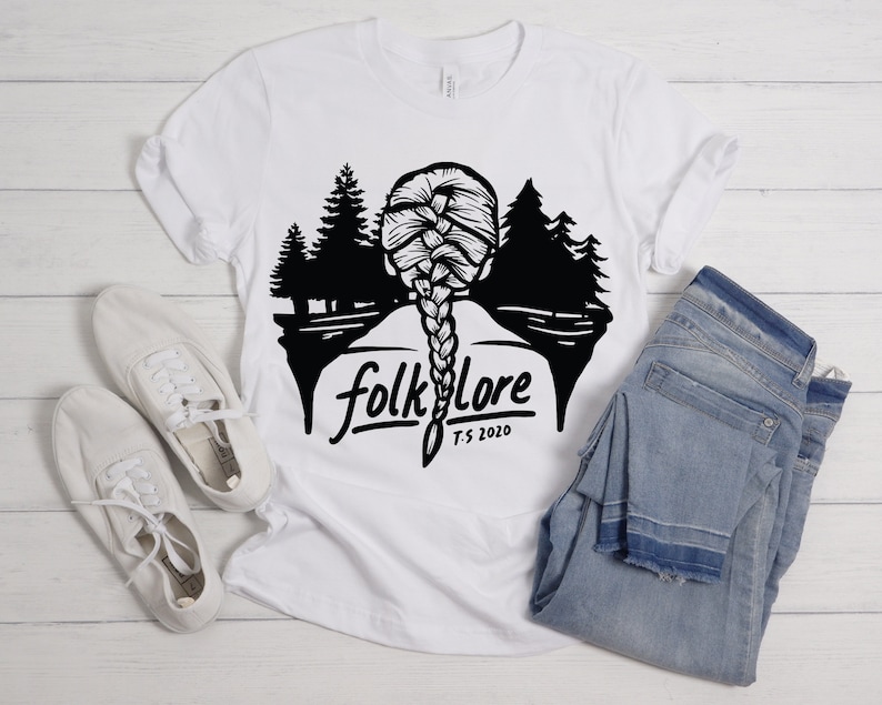 May include: White t-shirt with a black graphic featuring a braided hairstyle, trees, and the word "folklore" with "T.S. 2020" underneath. The shirt is paired with white sneakers and blue jeans.