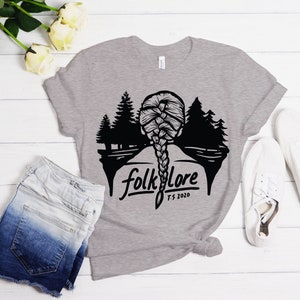 May include: A heather gray t-shirt featuring a black graphic of a braided hairstyle and a forest scene. The word "folklore" is printed below the graphic, with "T.S. 2020" underneath. The shirt is paired with denim shorts and white sneakers.