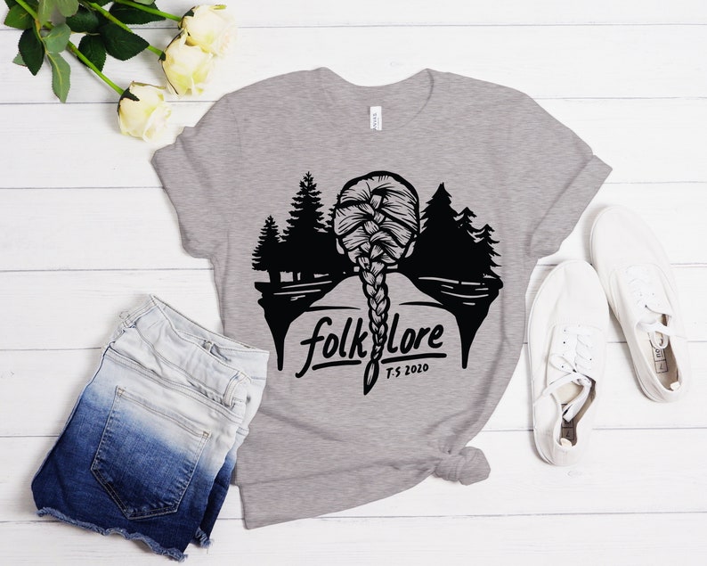 May include: A heather gray t-shirt featuring a black graphic of a braided hairstyle and a forest scene. The word "folklore" is printed below the graphic, with "T.S. 2020" underneath. The shirt is paired with denim shorts and white sneakers.