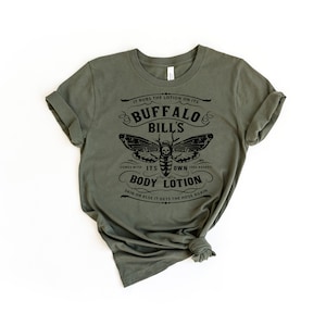 May include: A green t-shirt with a black graphic print of a moth with a skull and crossbones. The text on the shirt reads "It rubs the lotion on its Buffalo Bill's. Comes with its own body lotion. Skin or else it gets the hose again."