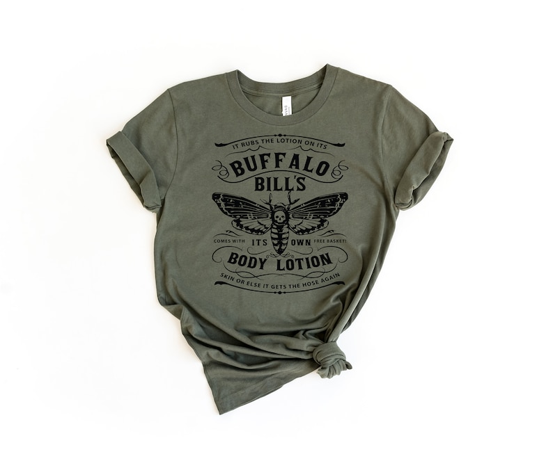May include: A green t-shirt with a black graphic print of a moth with a skull and crossbones. The text on the shirt reads "It rubs the lotion on its Buffalo Bill's. Comes with its own body lotion. Skin or else it gets the hose again."