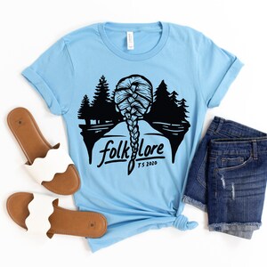 May include: Light blue t-shirt with a black graphic of a braided hairstyle, trees, and the word "folklore" with "T.S. 2020" underneath. The shirt is styled with denim shorts and white sandals.