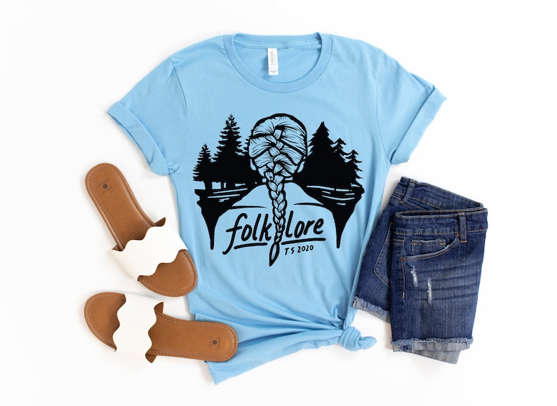May include: Light blue t-shirt with a black graphic of a braided hairstyle, trees, and the word "folklore" with "T.S. 2020" underneath. The shirt is styled with denim shorts and white sandals.