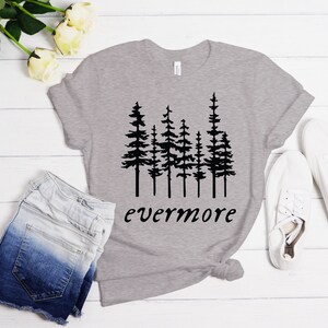 May include: A heather gray t-shirt with a black graphic of pine trees and the word "evermore" in a script font. The shirt is paired with denim shorts and white sneakers, suggesting a casual, outdoorsy theme.