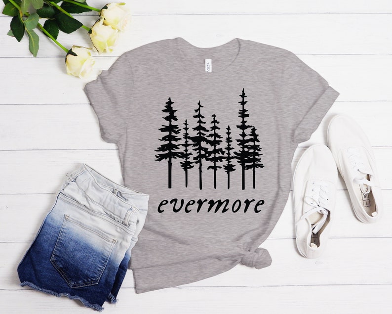 May include: A heather gray t-shirt with a black graphic of pine trees and the word "evermore" in a script font. The shirt is paired with denim shorts and white sneakers, suggesting a casual, outdoorsy theme.
