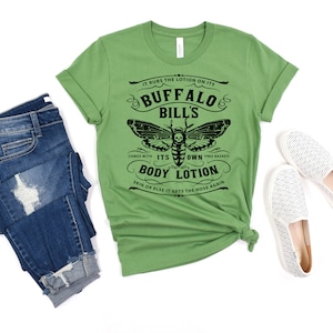 May include: Green t-shirt with a black and white graphic of a moth with a skull and crossbones. The text on the shirt reads "IT RUBS THE LOTION ON ITS BUFFALO BILL'S COMES WITH ITS OWN FREE BASKET BODY LOTION SKIN OR ELSE IT GETS THE HOSE AGAIN".
