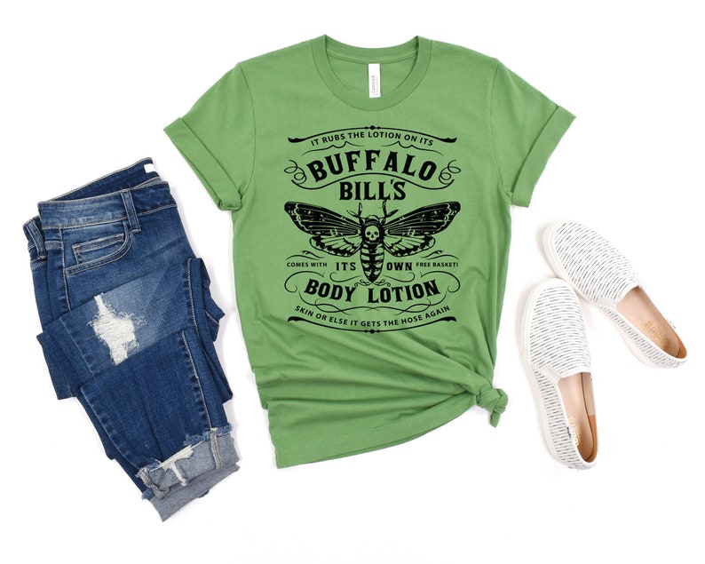 May include: Green t-shirt with a black and white graphic of a moth with a skull and crossbones. The text on the shirt reads "IT RUBS THE LOTION ON ITS BUFFALO BILL'S COMES WITH ITS OWN FREE BASKET BODY LOTION SKIN OR ELSE IT GETS THE HOSE AGAIN".