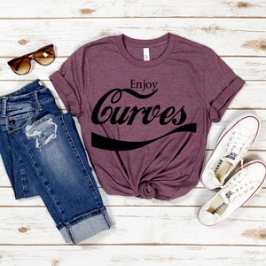 May include: A maroon t-shirt with the text "Enjoy Curves" in a black, stylized font. The shirt is laid out on a white wooden surface with a pair of blue jeans, sunglasses, and white sneakers.