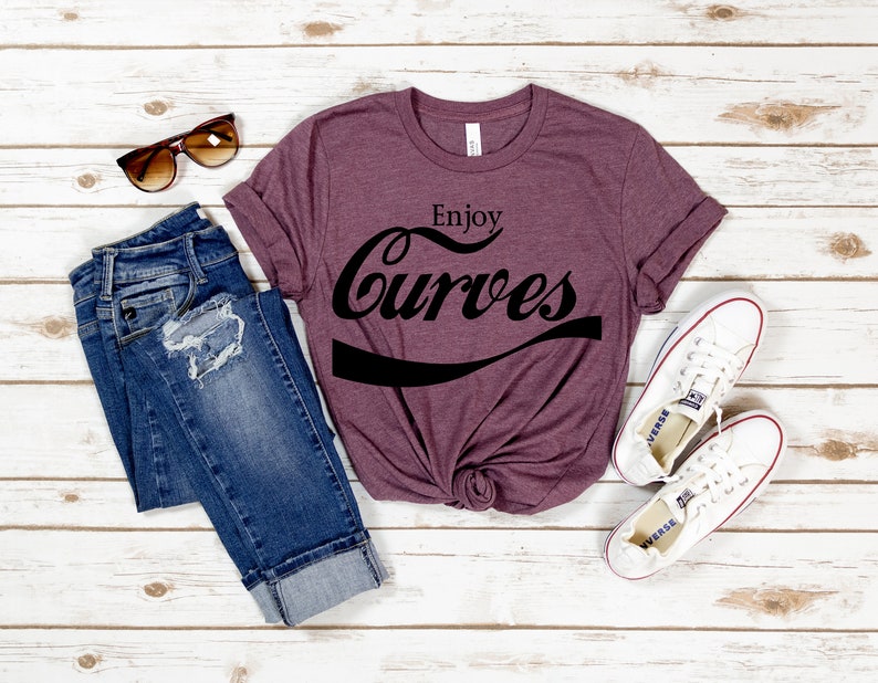 May include: A maroon t-shirt with the text "Enjoy Curves" in a black, stylized font. The shirt is laid out on a white wooden surface with a pair of blue jeans, sunglasses, and white sneakers.