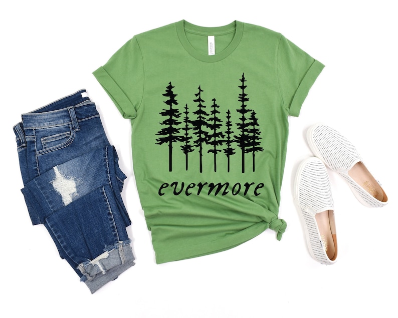 May include: A green t-shirt with a black tree graphic and the word "evermore" in black script. The shirt is paired with blue jeans and white slip-on shoes. The shirt is made of a soft material.