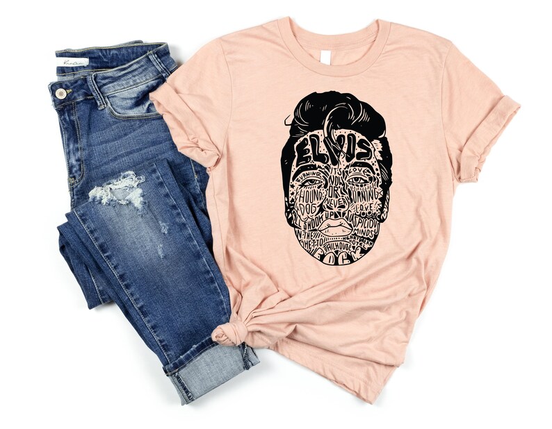 May include: A peach-colored t-shirt with a black graphic of Elvis Presley's face made up of words related to his music and career. The words include "Hound Dog", "Jailhouse Rock", "Love Me Tender", and "Suspicious Minds".