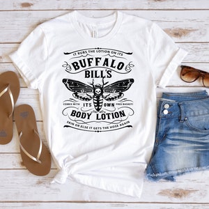 May include: White t-shirt with a black and white graphic design featuring a moth with a skull and crossbones. The text reads "It rubs the lotion on its Buffalo Bill's Comes with its own free basket! Body Lotion Skin or else it gets the hose again."