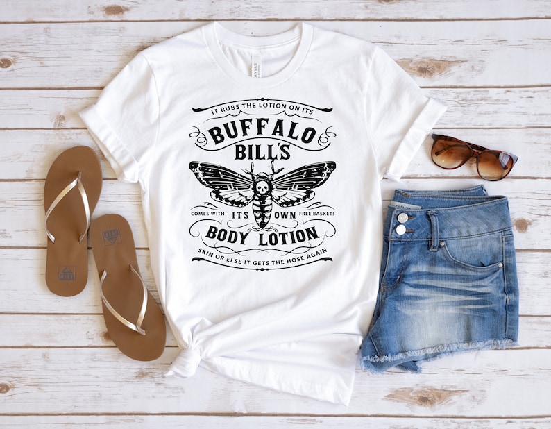 May include: White t-shirt with a black and white graphic design featuring a moth with a skull and crossbones. The text reads "It rubs the lotion on its Buffalo Bill's Comes with its own free basket! Body Lotion Skin or else it gets the hose again."